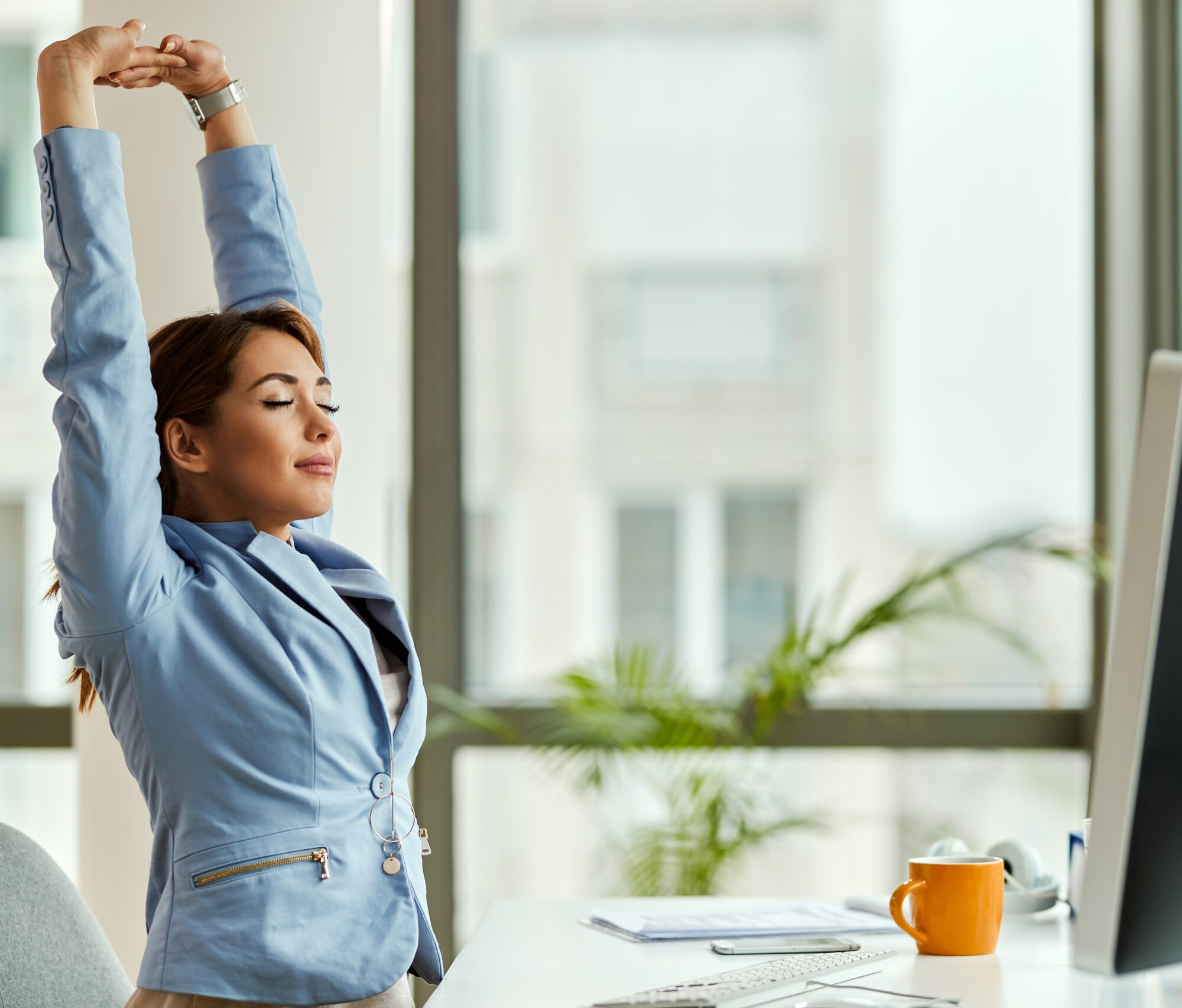 Smiling businesswoman with raised arms stretching at office desk Young smiling businesswoman stretching with her eyes closed while working on a computer in the office.